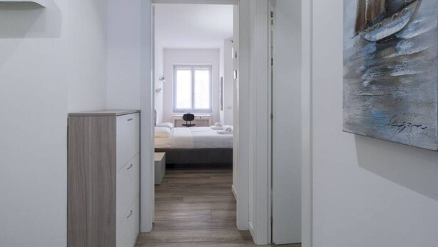 Un pasillo estrecho que conduce a un dormitorio con una cama, una mesita de noche y una ventana.