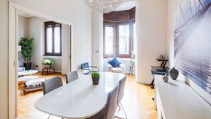 Ein moderner, gut beleuchteter Raum mit einem großen runden Tisch, bequemen Sitzmöglichkeiten, einer dekorativen Deckenleuchte und großen Fenstern, die für natürliches Licht sorgen.
