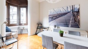 Ein modernes Wohnzimmer mit einem großen weißen Esstisch, grauen Stühlen, einem weißen Sofa, einem kleinen Beistelltisch und einem großen Fenster mit Blick auf Gebäude.