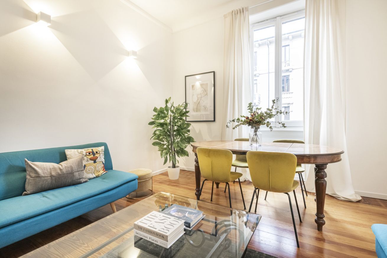 A modern living room with a blue sofa, a glass coffee table, a dining table with yellow chairs, and a potted plant.
