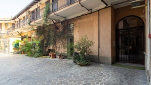 Une cour dallée de pierres, agrémentée de plantes en pot et bordée d’un bâtiment doté d’un portail noir et de balcons.