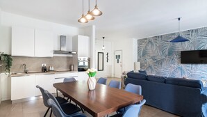 Uma cozinha moderna com armários brancos, uma mesa de jantar com cadeiras, um sofá azul e uma televisão fixada na parede.
