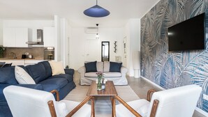 Uma sala de estar moderna com sofá, mesa de centro e cadeiras, uma cozinha ao fundo e uma televisão grande presa à parede.
