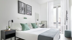 Quarto de hotel moderno com cama, mesa de cabeceira e escrivaninha com cadeira. Há duas fotos emolduradas na parede, um radiador e uma janela com cortinas.