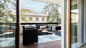 Varanda com mobiliário externo, um tapete estampado e vista para edifícios ao fundo.