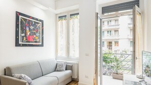 Ein modernes Wohnzimmer mit einem grauen Sofa, einem gerahmten abstrakten Gemälde, einem Heizkörper und Blick auf einen Balkon mit einem Blumentopf.