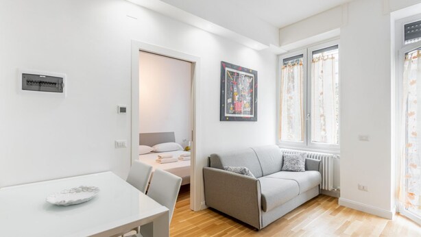 Ein moderner, gut beleuchteter Raum mit einem weißen Tisch, einem grauen Sofa und einem Bett mit weißem Kopfteil.