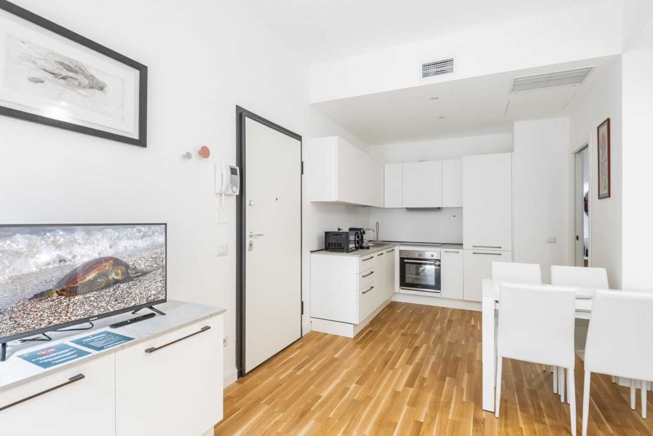A modern kitchen with white cabinets, a dining area with white chairs, and a television displaying a turtle on a beach.