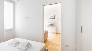 Ein modernes Schlafzimmer mit einem ordentlich bezogenen Bett, Holzboden und Blick in einen weiteren Raum mit Schreibtisch und Stuhl.