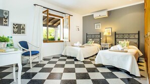 Une chambre d’hôtel avec deux lits, un bureau et une chaise. Il y a une fenêtre avec des rideaux, un climatiseur fixé au mur et des œuvres d’art encadrées accrochées au mur.