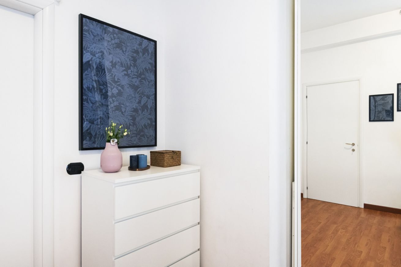 A hallway with a white dresser, a framed artwork, a vase with flowers, and a door in the background.