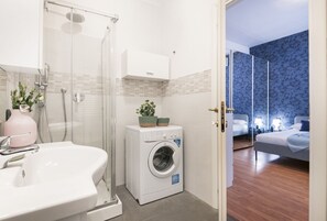 Een compacte badkamer met een wastafel, douche en wasmachine. Een slaapkamer met een bed en nachtkastjes is door een geopende deur te zien.
