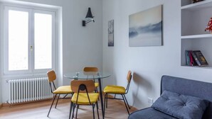 Una habitación con una mesa de comedor y sillas, un sofá, un radiador y una estantería llena de libros y objetos decorativos.