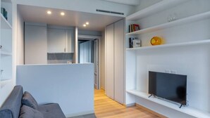 Una sala de estar moderna con un sofá gris, un televisor de pantalla plana sobre un soporte blanco y una estantería con diversos objetos.