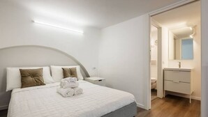Une chambre d’hôtel avec un lit bien fait, une table de chevet, une petite commode, une salle de bain avec un miroir et un plancher en bois.