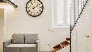 Une pièce avec un canapé gris, une horloge murale, une fenêtre et un escalier.