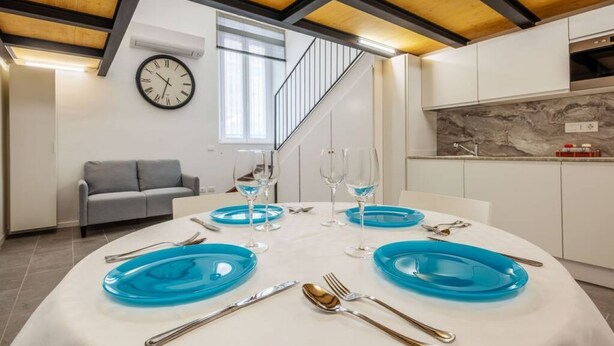 Un espace de salle à manger moderne avec une table ronde pour quatre personnes, agrémentée d’assiettes bleues et de couverts en argent. Un canapé gris est placé contre le mur, et une horloge est fixée au-dessus.