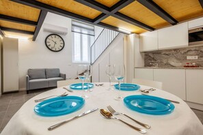 Un espace de salle à manger moderne avec une table ronde pour quatre personnes, agrémentée d’assiettes bleues et de couverts en argent. Un canapé gris est placé contre le mur, et une horloge est fixée au-dessus.