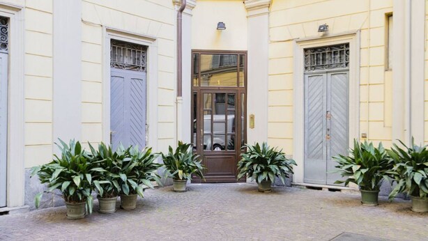 Une ruelle étroite pavée de pierres, avec une porte centrale, flanquée de deux grandes portes grises, et des plantes en pot de chaque côté.