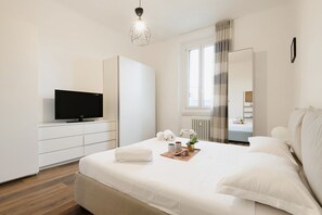 Een moderne slaapkamer met een bed, een televisie op een witte kast, een raam met gordijnen en een radiator.