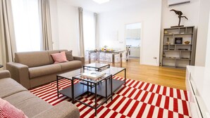 Een moderne woonkamer met een bank, salontafel en een gestreept vloerkleed. Achtergrond toont een keuken met een eethoek.