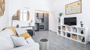 Un salon moderne avec un canapé gris, un téléviseur posé sur un meuble blanc, et une cuisine en arrière-plan.