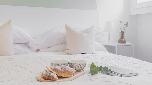 Una cama bien hecha con almohadas, una mesita de noche con una lámpara y una planta, y una bandeja con pasteles y un libro.
