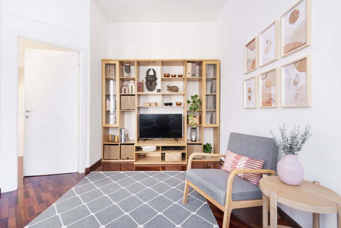 Una sala de estar con televisor, estanterías de madera, alfombra gris, sillón gris y una mesita con un florero.