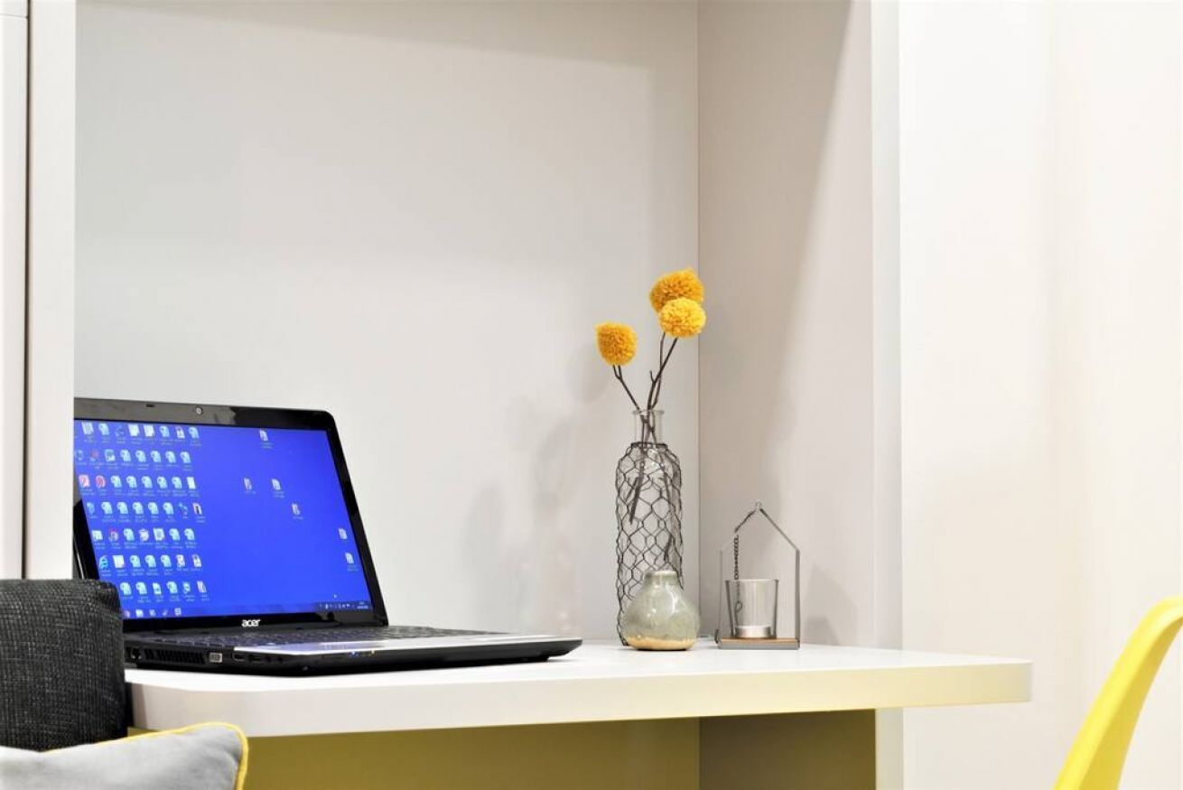 A laptop on a white desk with a vase of yellow flowers and a glass candle holder.