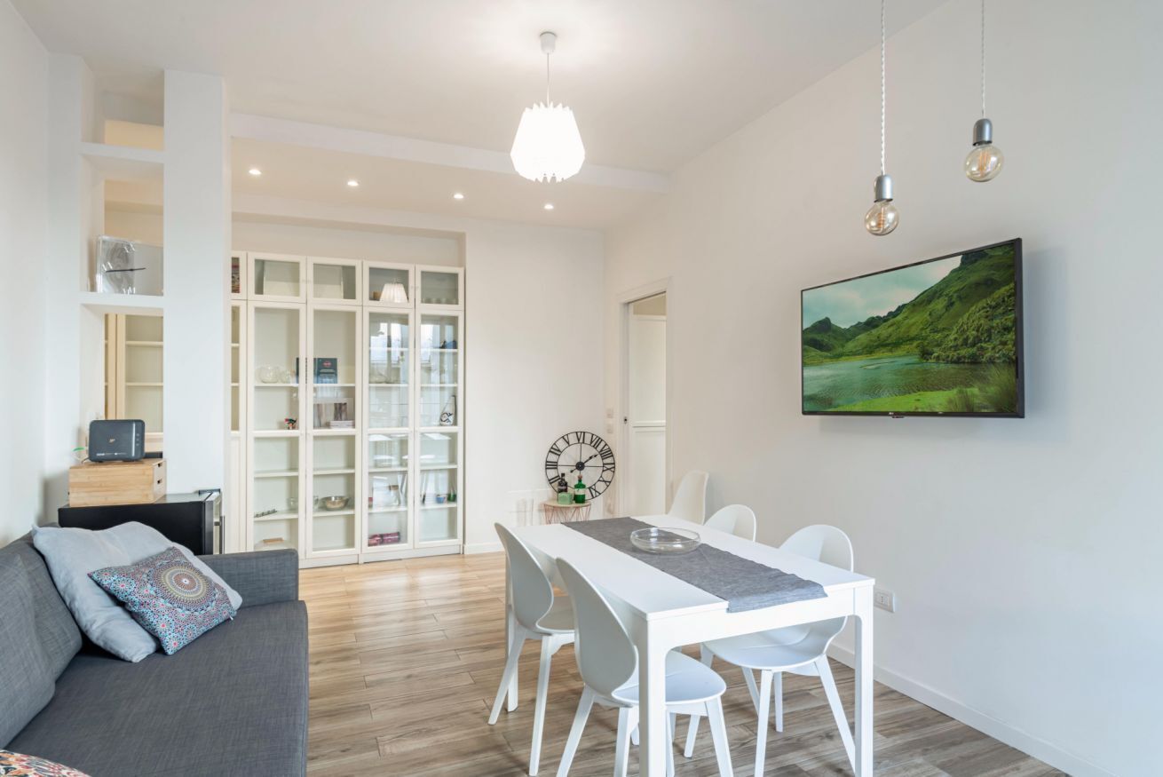 Uma sala de estar moderna com área de jantar, mobiliário branco, um relógio grande e uma TV na parede.