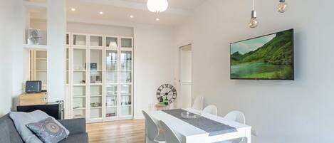 Uma sala de estar moderna com área de jantar, mobiliário branco, um relógio grande e uma TV na parede.