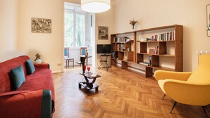 Un salon avec un canapé rouge, un fauteuil jaune, une bibliothèque en bois, un téléviseur et un parquet à motifs chevrons.