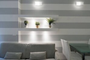 A modern living room with a grey sofa, a glass dining table, and three shelves with plants.