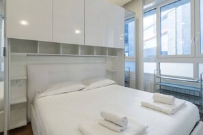 A modern hotel room with a large bed, white linens, and a view of the city through large windows.