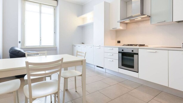 A modern kitchen with white cabinetry, a built-in oven, and a dining area with a table and chairs. - Easylife - Spacious and new apt in Città Studi (Milano)