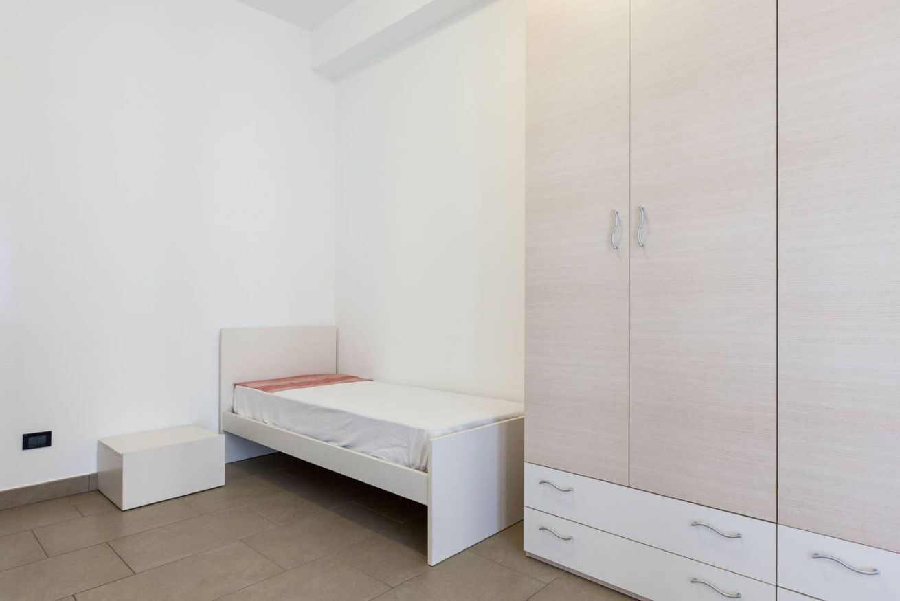 A small, white bedroom with a single bed, a white wardrobe with multiple drawers, and a small white bench.
