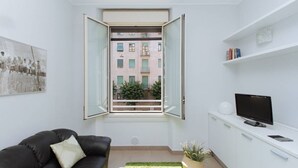 A modern living room with a black leather sofa, a television on a white cabinet, and a window with an open view of a building. - Easylife - Spacious and new apt in Città Studi (Milano)