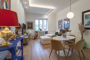 Un salon moderne avec une table de salle à manger ronde, des canapés beige et une console bleue surmontée d’une lampe rouge.