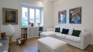 Ein modernes Wohnzimmer mit einem weißen Ecksofa, einem Couchtisch mit Glasplatte, einem Bücherregal und zwei abstrakten Bildern an der Wand.