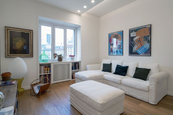 Un salon moderne avec un canapé d’angle blanc, une table basse en verre, une bibliothèque et deux tableaux abstraits accrochés au mur.
