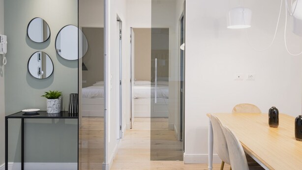 A modern hotel room with a glass partition separating the living area from the bedroom. There is a wooden dining table with chairs, a small black side table with a plant, and a round mirror on the wall. - Easylife - Precious Pearl in Porta Romana (Milano)