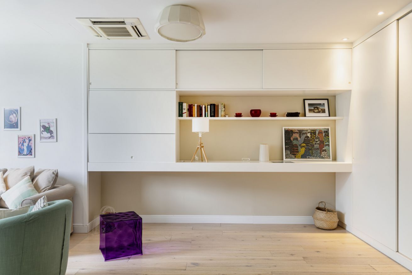 Una sala de estar moderna con un sofá, un baúl morado, una estantería con libros y adornos, y piso de madera.