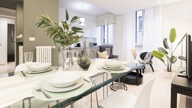 Un moderno comedor con una mesa de vidrio preparada para cuatro, rodeada de sillas blancas. Una planta grande está en el centro y la habitación tiene una paleta de colores neutros, con paredes blancas y una pared de acento gris.