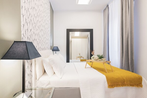 Habitación de hotel con cama, mesita de noche con lámpara, espejo y una pequeña mesa con bandeja.