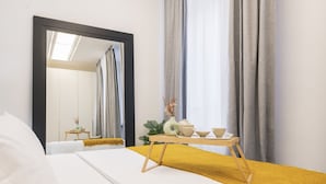 Una habitación de hotel con una cama, una mesita de noche con un florero y cuencos, un espejo y cortinas.