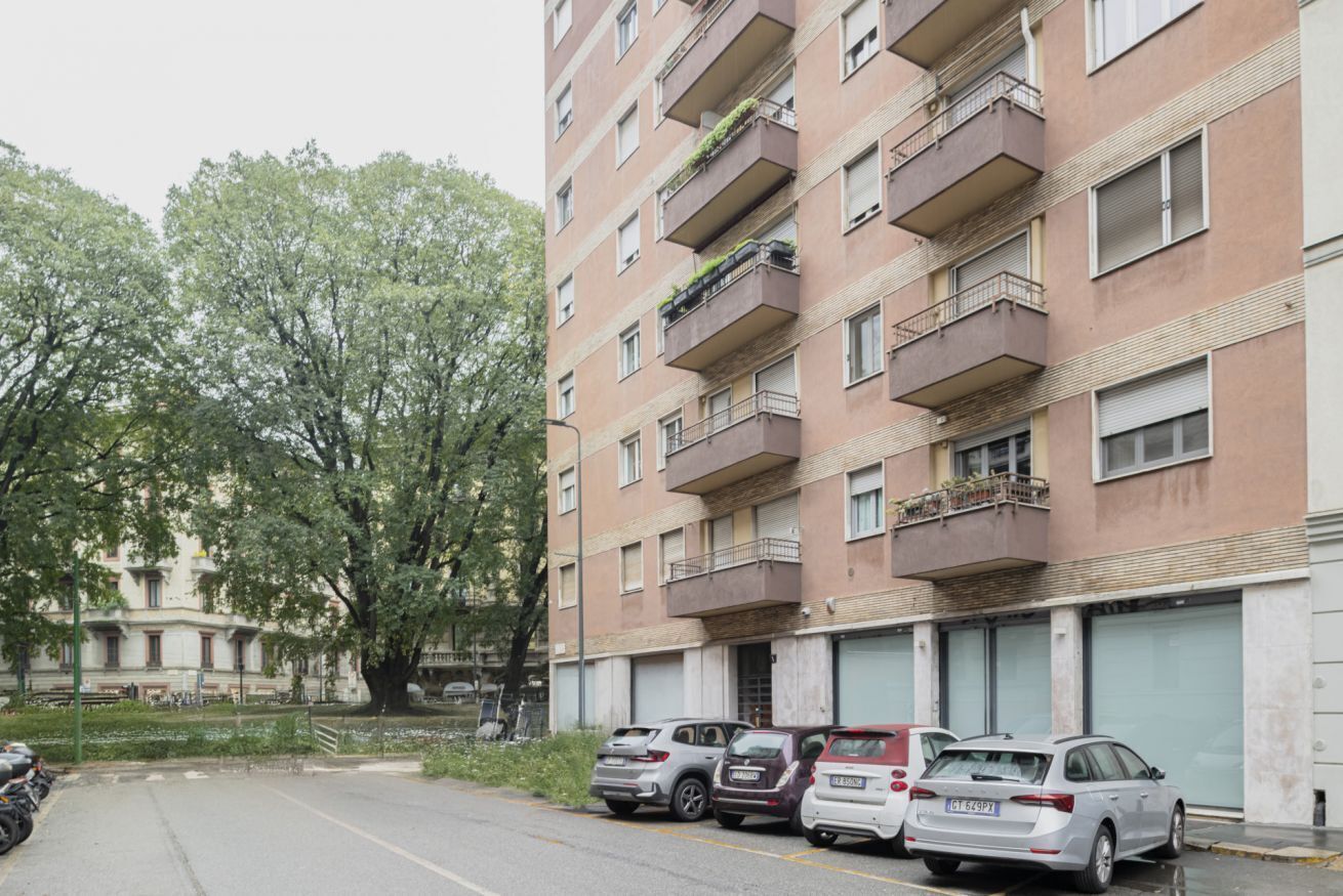 A multi-story residential building with balconies, parked cars, and a tree in the foreground.