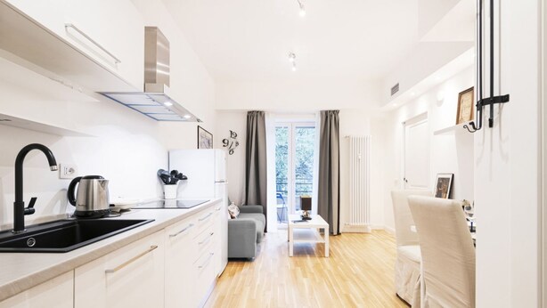 A modern kitchen with white cabinets, a black sink, and a kettle. There is a grey sofa and a white table in the living area. - Easylife - Bright and charming flat with balcony (Milano)
