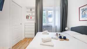 A modern bedroom with a large bed, a bedside table with a tray, a window with curtains, a white wardrobe, and a shelf with books and decorative items. - Easylife - Bright and charming flat with balcony (Milano)