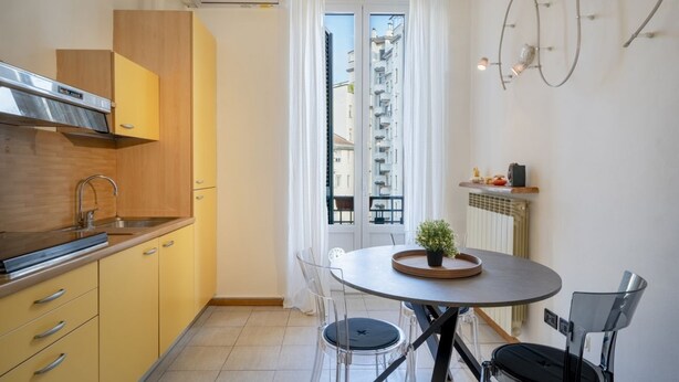 A modern kitchen with yellow cabinets, a stainless steel range hood, a round dining table with chairs, and a view of a building through a window.