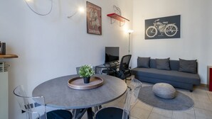A modern living room with a grey sofa, a round dining table with chairs, a mounted TV, and a framed picture of a motorcycle.